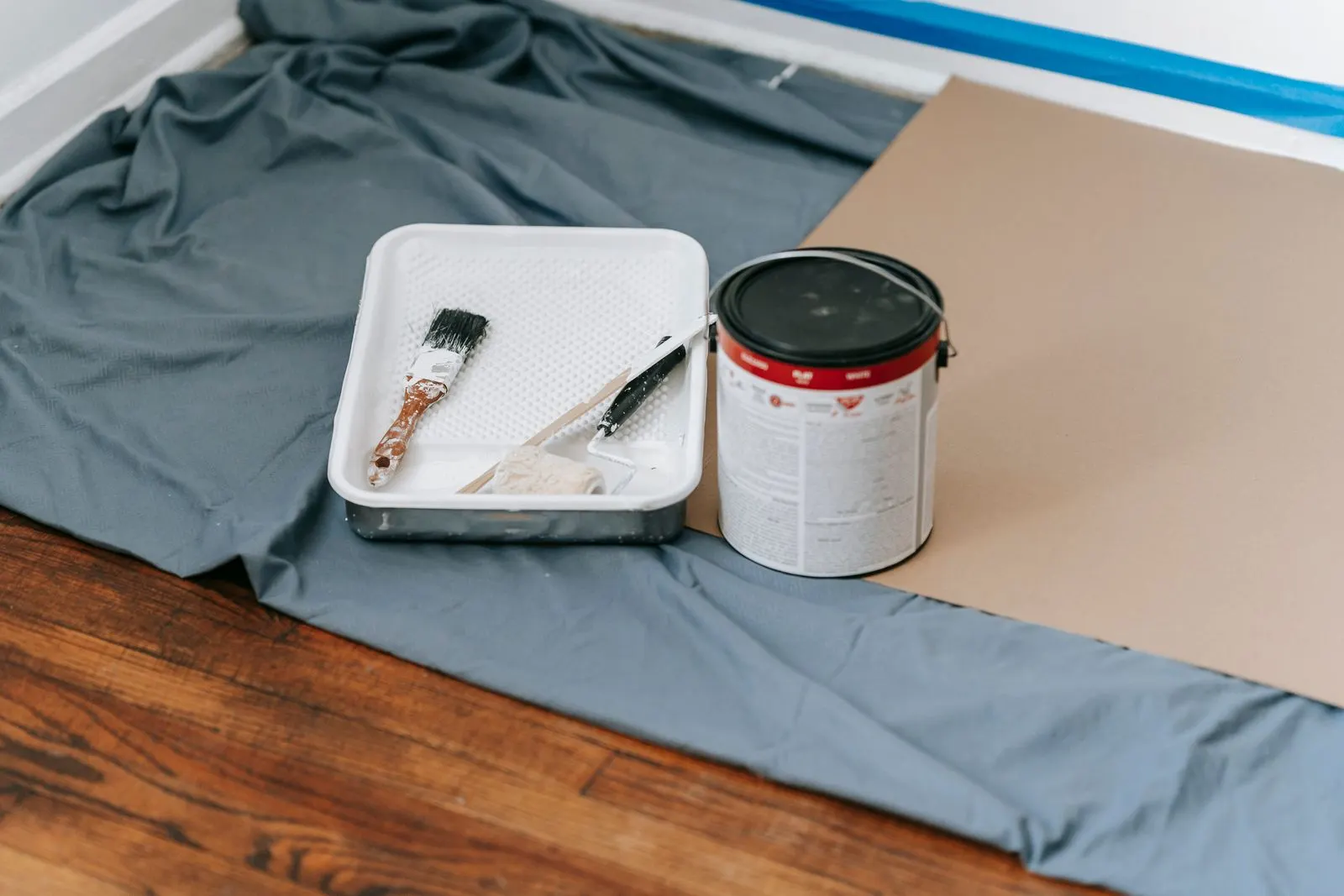Paint tray holding a brush and roller next to a paint can on a gray drop cloth over a wood floor