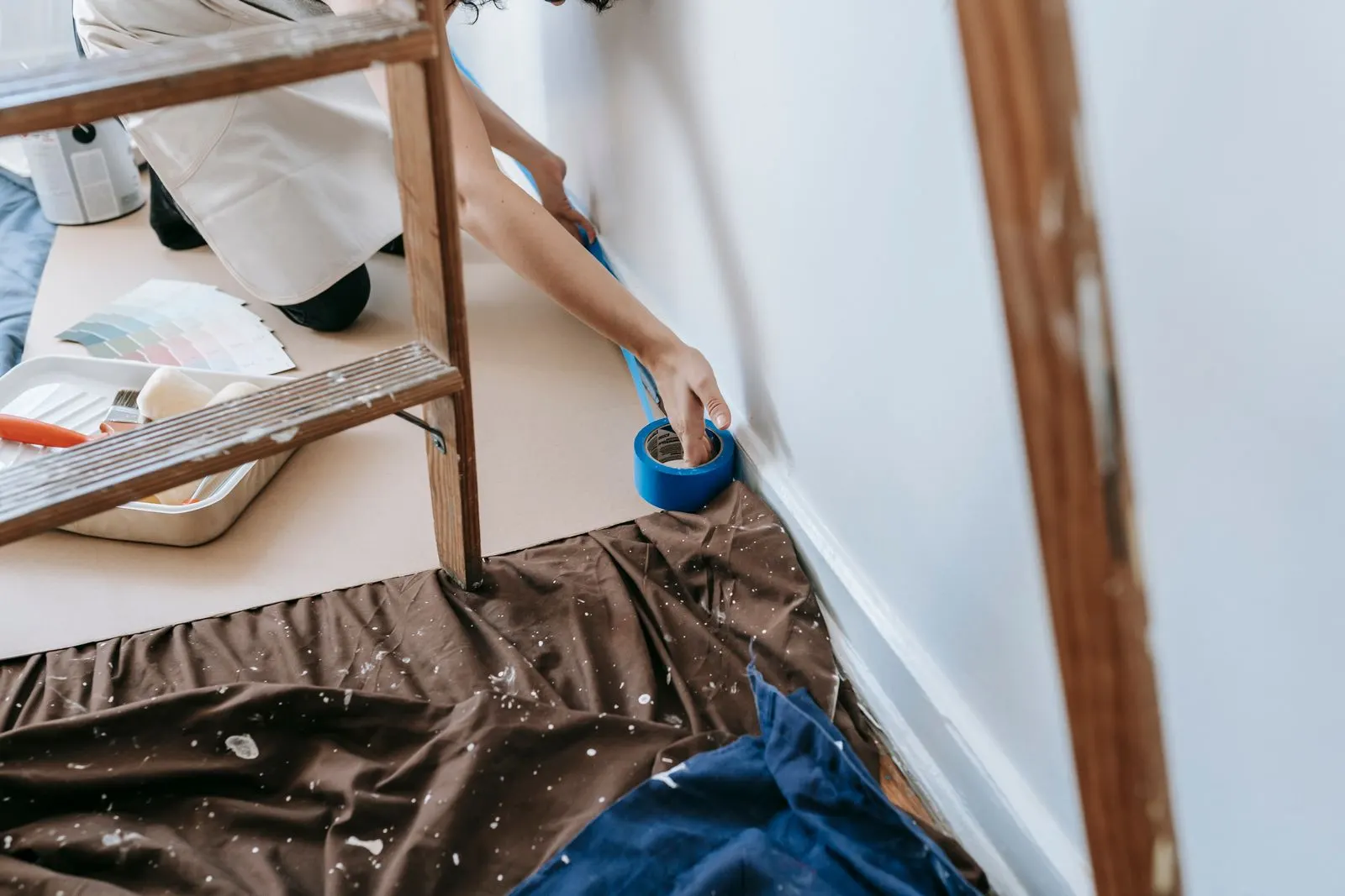 Kneeling person reaching for a roll of blue painter's tape at the base of a wall, with drop cloths, a paint tray and color swatches nearby