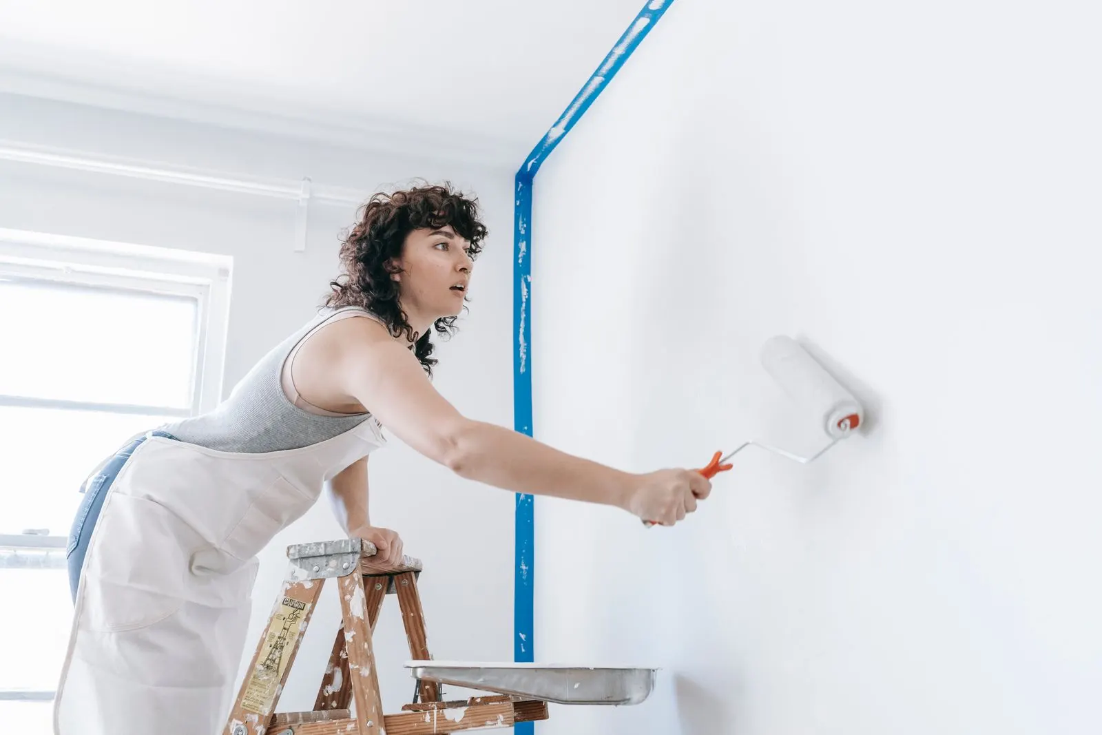 Painter in white overalls leaning on a stepladder while rolling white paint onto an interior wall beside a blue tape line